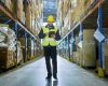 Auditor Wearing Hard Hat with Tablet Computer Counts Merchandise in Warehouse. He Walks Through Rows of Storage Racks with Merchandise.